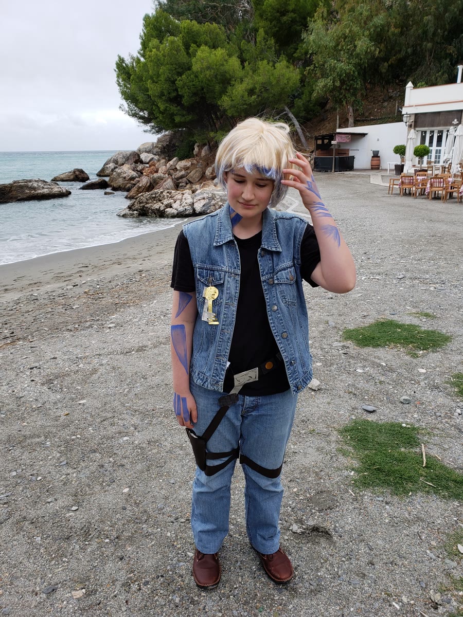 A cosplayer with a blonde wig streaked with blue, blue body paint markings along both arms, and a key pendant necklace. They wear a sleeveless denim vest over a black t-shirt, blue jeans, and brown shoes, with a black strap across the body. One hand is raised near their head as they pose on a gravelly shoreline, the sea and a building behind them.