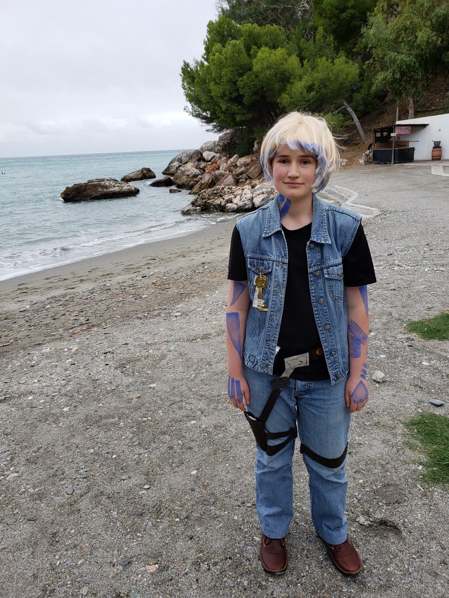 The same cosplayer standing and facing the camera on a pebbly beach. They wear a blonde wig streaked with blue, blue body paint markings on their arms, a key pendant necklace, a sleeveless denim vest over a black t-shirt, blue jeans, and brown shoes, with a black strap across the body. The sea, rocks, and a forested headland rise behind them under an overcast sky.