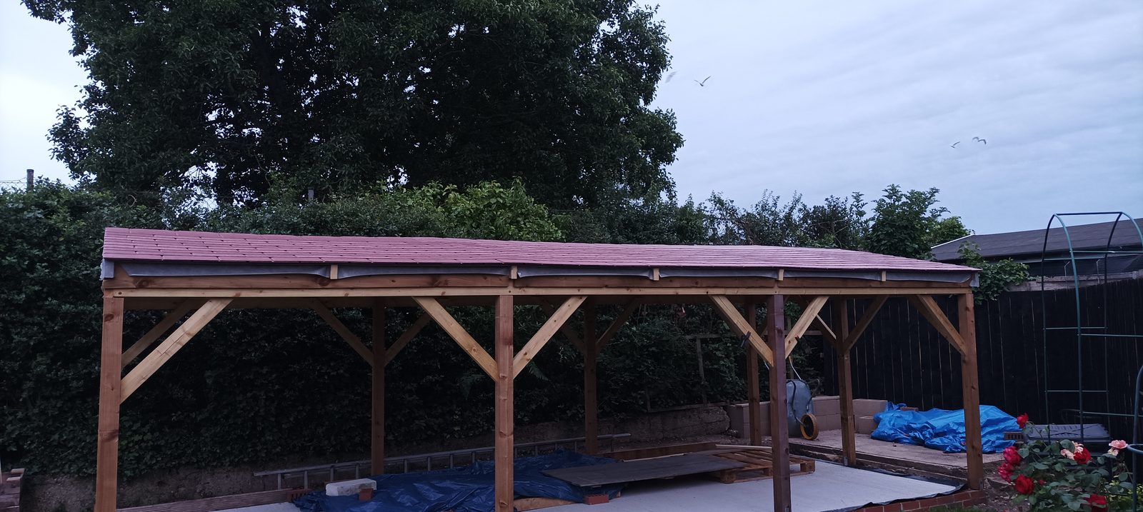 The fourth Workshopshed under construction: a large timber-framed structure with a freshly tiled red roof and open sides, standing on a concrete base in a garden bordered by trees and hedges.