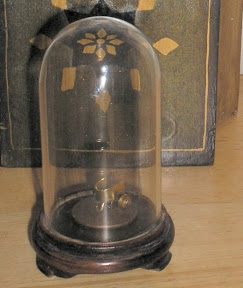 A small kinetic sculpture under a glass dome on a turned wooden base, displayed in front of an antique book.