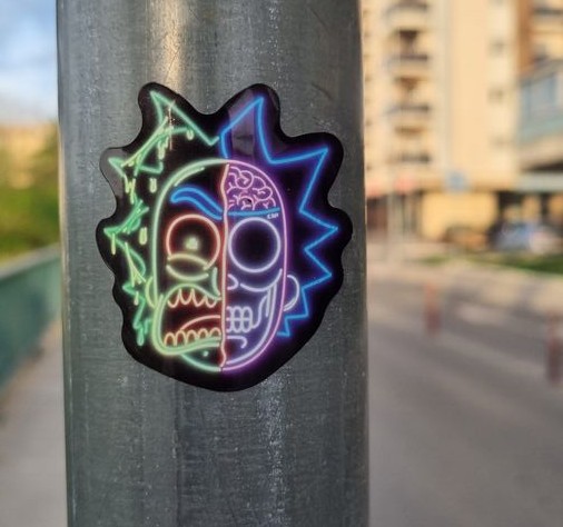 A neon-colored sticker on a metal railing showing a cross-section of Rick Sanchez's head from Rick and Morty, with visible brain, eye, and mouth details in glowing blue, purple, red, and green lines. Buildings visible in the blurred background.